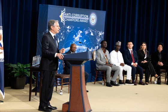Secretary Blinken speaks at a lectern. A poster behind him says, “Anti-Corruption Champions Award,” and award winners are sitting to his left