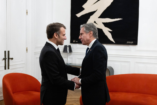 Secretary Blinken and French President Emmanuel Macron shake hands and smile.