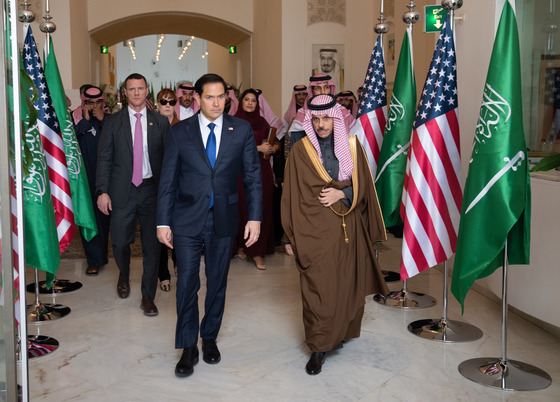 Secretary Marco Rubio and Saudi Arabian Foreign Minister Faisal bin Farhan walking in front of a group down a hallway lined with flags.