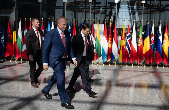 Secretary Rubio, U.S. Ambassador to NATO Matthew Whitaker, and two other men walking in front of a row of flags