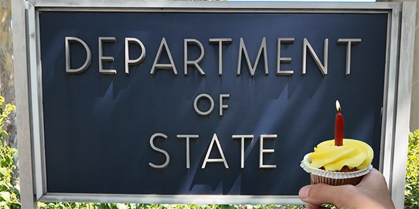 Photo of someone holding a cupcake with a lit candle in front of a signage that reads Department of State