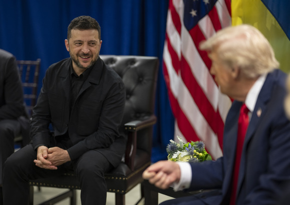President Trump is seated next to Ukrainian President Volodymyr Zelenskyy with the U.S. and Ukraine flag behind them.