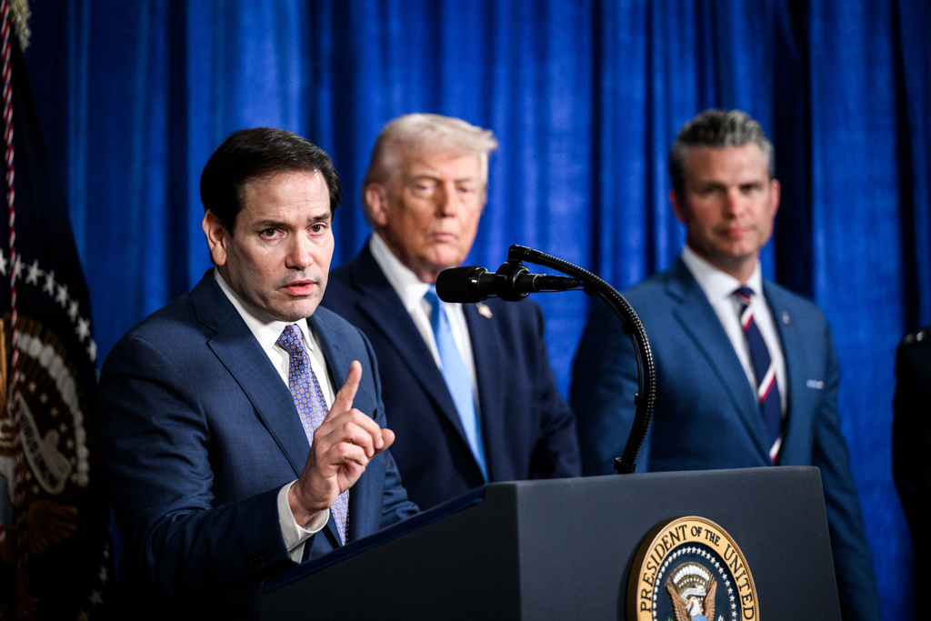  Secretary Rubio standing at a lectern speaking with one finger raised and President Trump standing behind him.