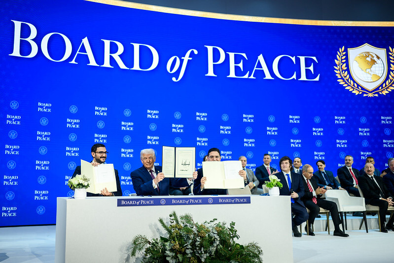 President Trump sits behind a desk among other world leaders holding up a signed charter for the Board of Peace.