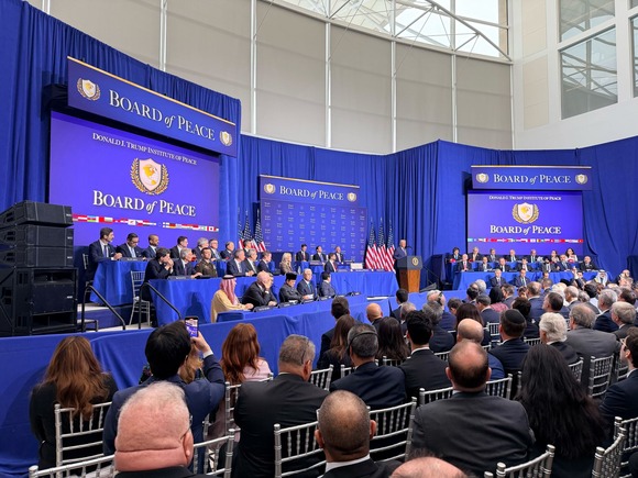  President Trump gives remarks on a stage in front of an audience with “Board of Peace” screens behind him.