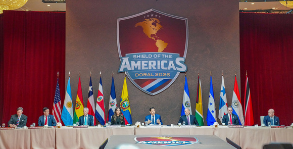 A speaker sits in the center of a table, in front of national flags flanking a shield-shaped sign that reads “Shield of the Americas, Doral 2026.” 