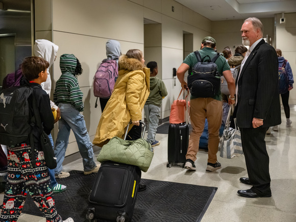 An official in a suit stands to the right inside an airport corridor as he greets casually dressed travelers, who are arriving from a flight. 