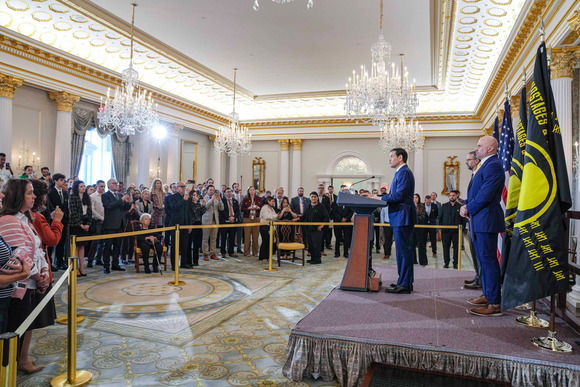 A speaker in a gilded ceremonial room stands at a podium to the right of his audience on a raised platform in front of deputies and flags.