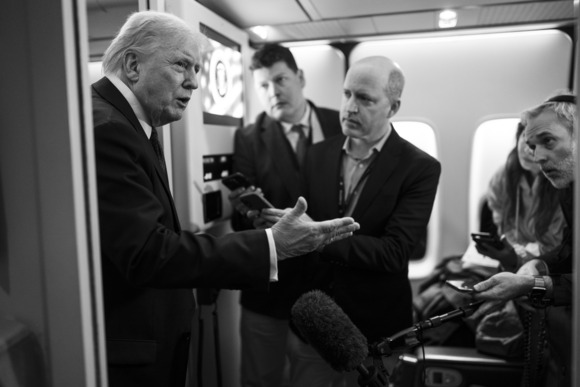 President Trump, pictured to the left inside a plane, gestures towards four journalists, who hold out devices to record his comments.