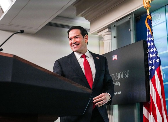 Secretary Rubio smiles behind a lectern, with a U.S. flag in the background.
