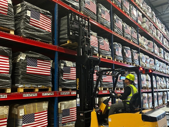 A man in fluorescent clothing and a hard hat sits in a forklift and moves boxes marked with U.S. flags.  