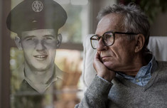 older man gazing out window with image of his younger self in military uniform