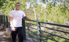 Man in t-shirt and jeans standing outdoors along a bridge.