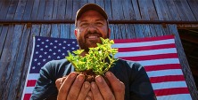 Man holding a small plant with American flag in the background.