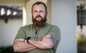 Veteran in olive green tee shirt with his tatooed arms folded across his chest.