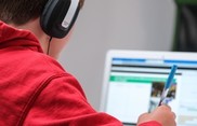 student from behind wearing over the ear headphones, while writing and looking at a laptop computer