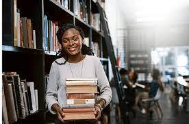 female college student standing in library stack area while holding a pile of books