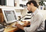 Side view of a man sitting down while looking at a laptop and holding an office phone to his ear with his shoulders