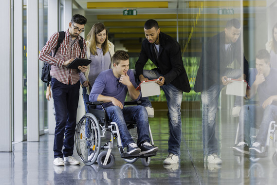 Three college students stand while one sits in a wheel chair, in a hallway