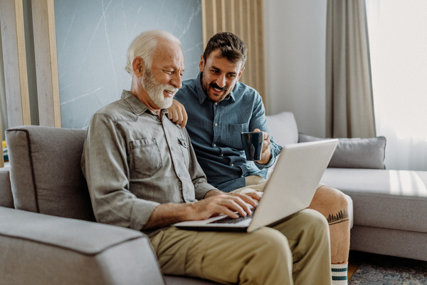A younger man helps an older man navigate a laptop computer while sitting together on a couch.