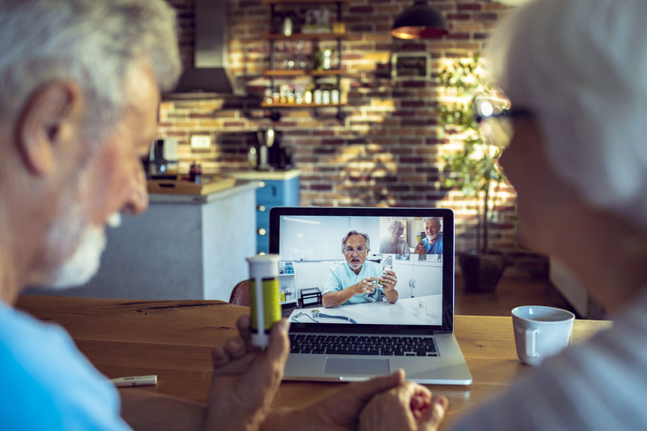 Two older individuals are meeting with a doctor over the computer. One is holding a pill bottle.