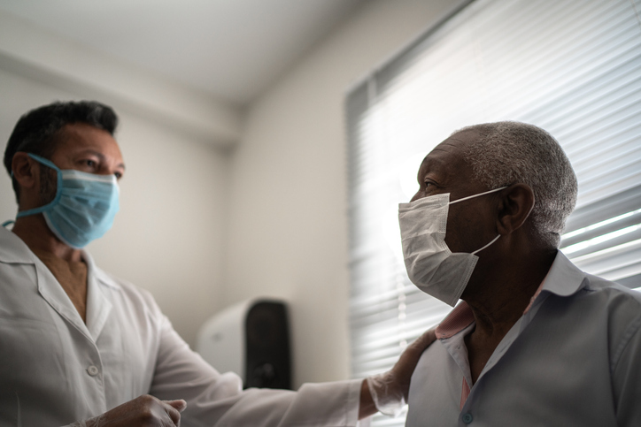 A masked doctor places his hand on a masked male patient's shoulder