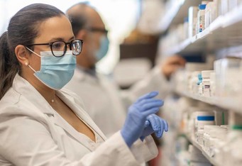 A pharmacist wearing a mask searches for medication on shelf