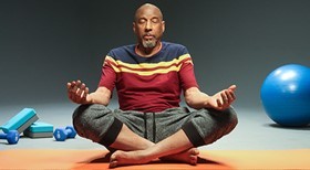 A man sits crossed-legged on the floor, palms up and eyes closed meditating. He is surrounded by an exercise ball and hand weights