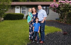 Picture of a family comprising of mom, dad, daughter and son smiling in front of a house. 