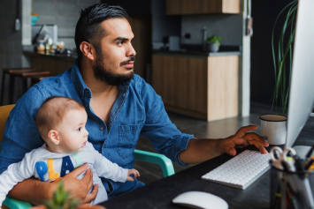 A man types on a computer keyboard with a baby in their lap.