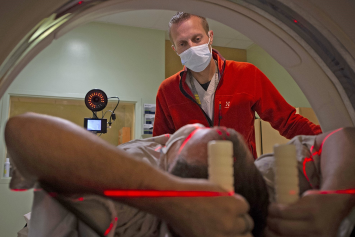 A patient lies inside a PET scan machine with a red bar of light crossing his body, as a medical technician stands outside and operates the machine.