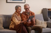 An older man and woman sit on a couch, smiling and looking at a tablet. 