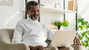 Older man sits on a couch, and smiles at his laptop. 