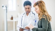 Doctor in white coat with stethoscope consulting a patient holding a clipboard in a bright medical office.