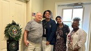 Four people standing together in a hallway near a door decorated with a United States Army wreath and emblem.
