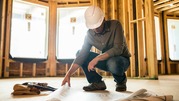 Construction worker wearing a white hard hat reviewing blueprints spread on the floor next to a tool belt.
