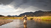 A man standing in the middle of a river casting a fly fishing line with mountains and cloudy sky in the background.