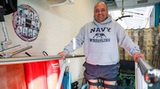 Veteran Edward Afanador wearing a Navy Wrestling hoodie using parallel bars for walking support in a rehabilitation setting.