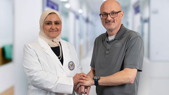 A female doctor in a white coat is holding the hand of a male patient.