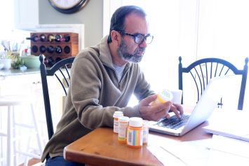 Man holds a prescription bottle in one hand and types on laptop with the other.