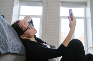 Woman sitting on floor in front of a bed looks up at a mobile phone in her hand.