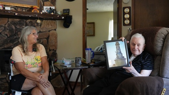 Ron Gasper sitting in a recliner holding a framed photo, while a woman sits nearby in a living room with stone fireplace.