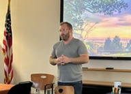person in grey t-shirt standing in front of conference room with a colorful background