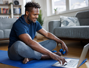 Black male sitting on yoga mat browsing on a laptop.