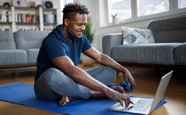 Black male sitting on yoga mat browsing on a laptop.