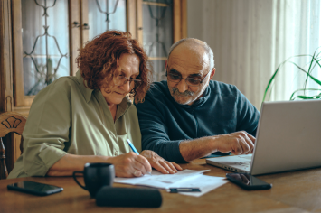 A Veteran writes notes down on a sheet of paper while her husband types on a laptop