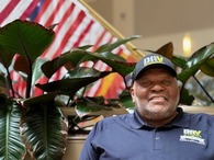 a man wearing a DAV baseball hat sitting in front of flags