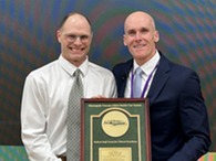 two people wearing shirts with ties holding an award in front of a big screen