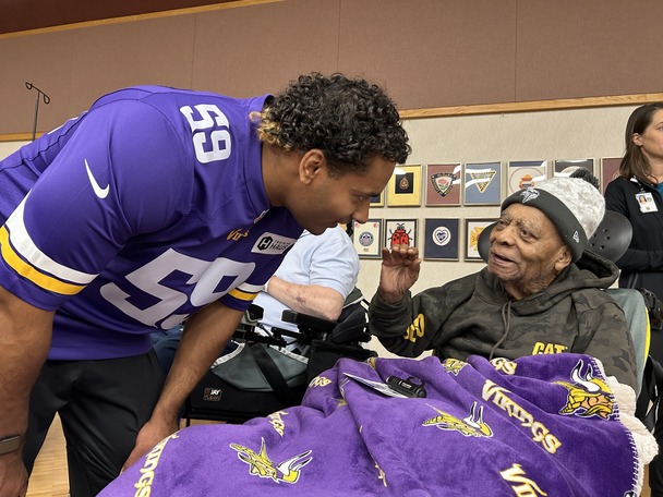 A football player wearing a purple jersey leaning over and talking to a person in wheelchair with a matching purple color blanket