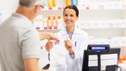 Pharmacist handing a prescription bag to an elderly male customer at the pharmacy counter.